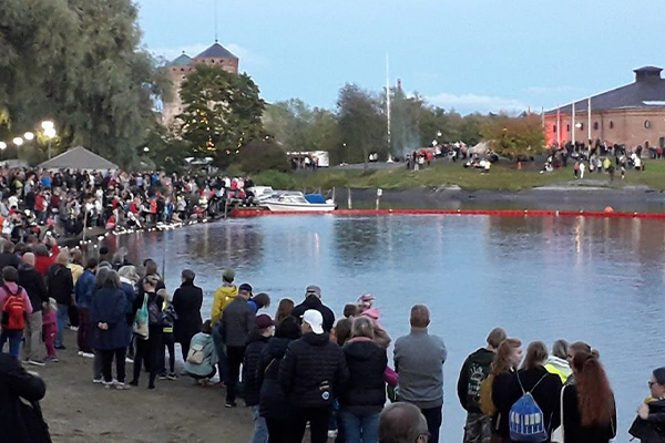Syksyinen kuva, jossa runsaasti ihmisiä osallistuu rantatapahtumaan. Taustalla näkyy Olavinlinna.