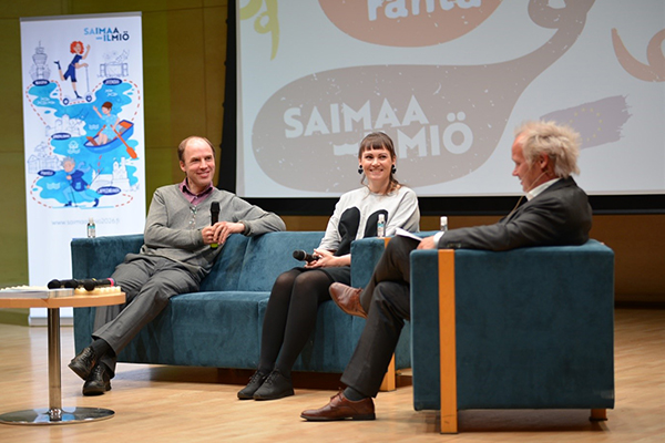 Hankekoordinaattorit Reko Tammi ja Anu-Anette Varho istuvat sohvalla, Jani Halme haastattelee. Kuvassa näkyy Saimaa-ilmiön mainos.